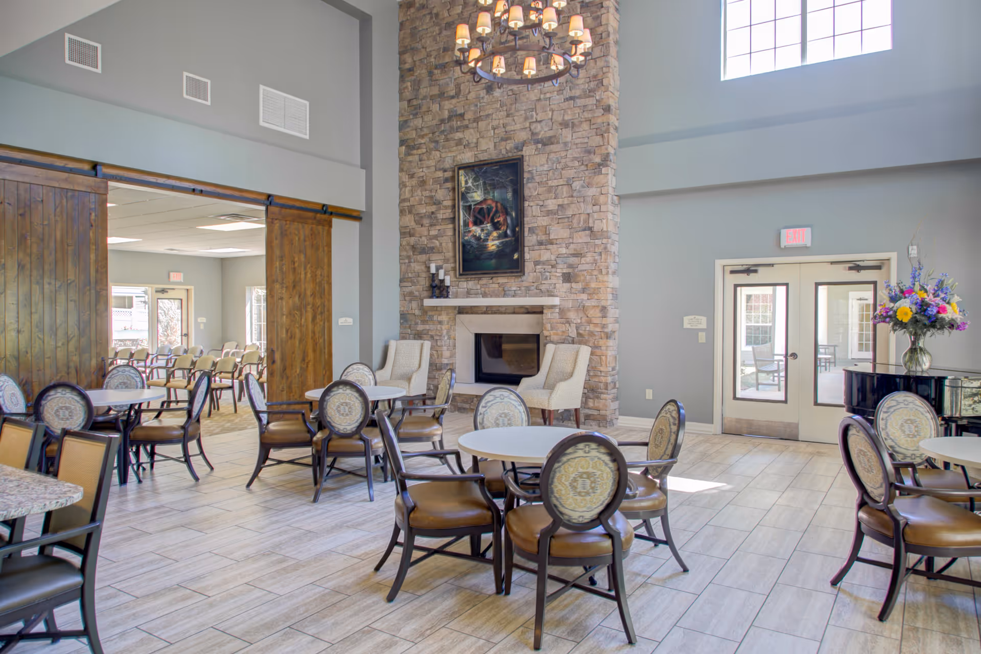 A bright and spacious common area with round tables and chairs arranged for seating. The room features a tall stone fireplace with a painting above it, two beige armchairs, large windows letting in natural light, a chandelier, and a vase of colorful flowers on a black piano near the double glass doors leading outside.
