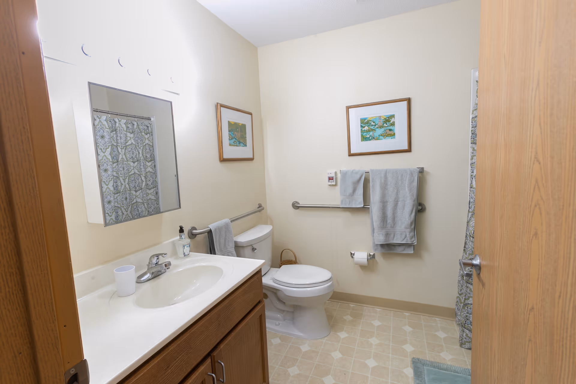 A clean bathroom with a white sink and countertop, wooden cabinet, a mirror above the sink, a toilet with grab bars on the wall, two framed pictures, a towel rack with two gray towels, a patterned shower curtain, and a light-colored tiled floor.
