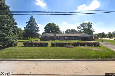 Single-story brick building with a chimney, surrounded by green lawn, bushes, and trees under a partly cloudy blue sky. A paved driveway is visible on the right side of the building.