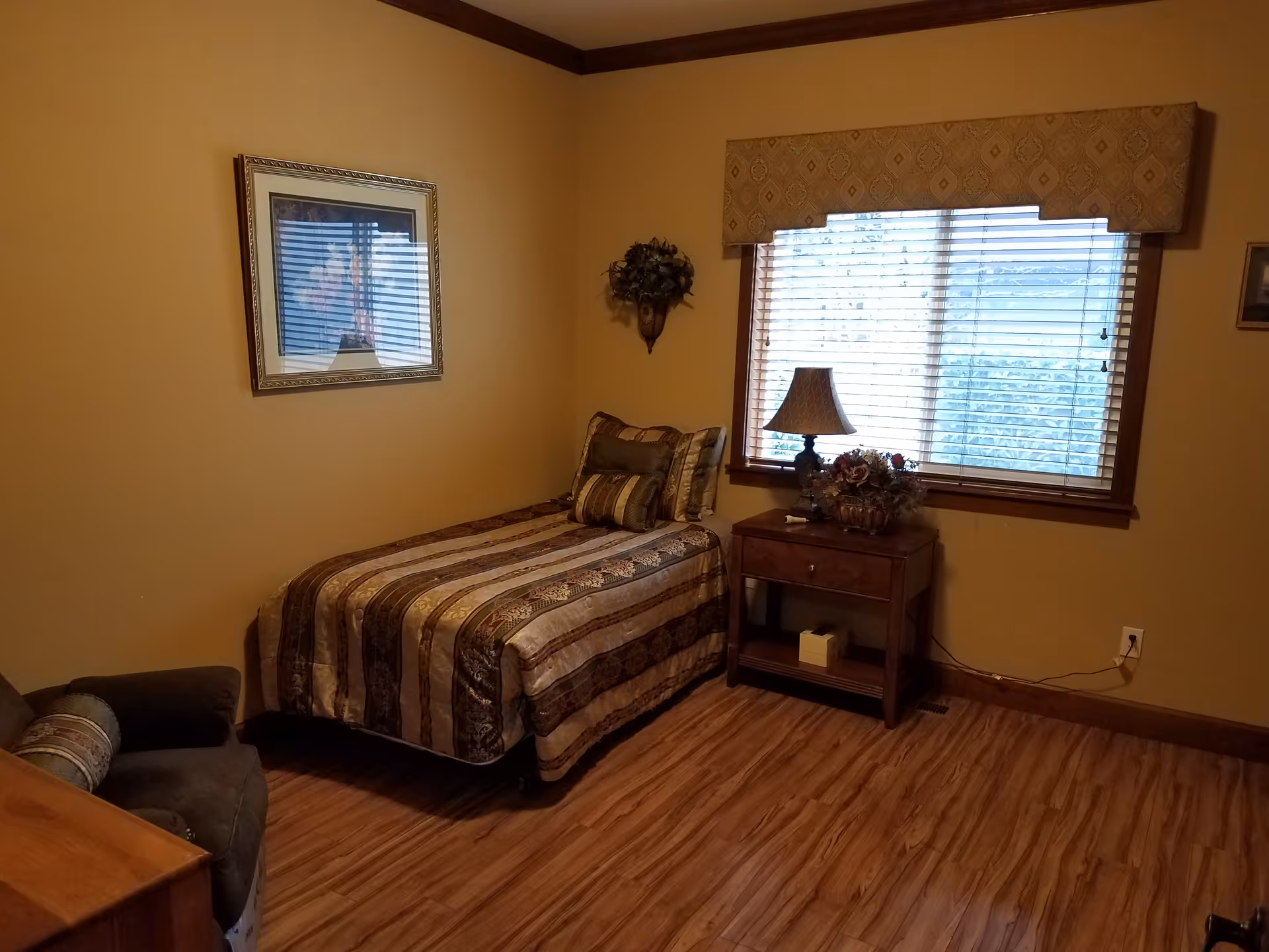 A cozy bedroom with a single bed covered in a striped brown and beige comforter. There are several pillows on the bed. Next to the bed is a wooden nightstand with a lamp and a floral arrangement. A window with blinds and a decorative valance lets in natural light. The room has wooden flooring, beige walls, a framed picture on the wall, and a comfortable armchair in the corner.