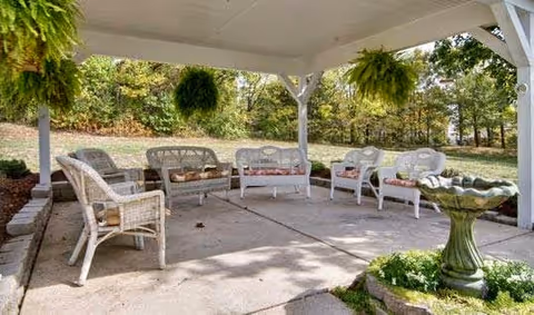 Outdoor covered patio area with white wicker chairs and loveseats arranged in a circle on a concrete floor, surrounded by hanging green ferns and a birdbath in the foreground, with trees and grass in the background.