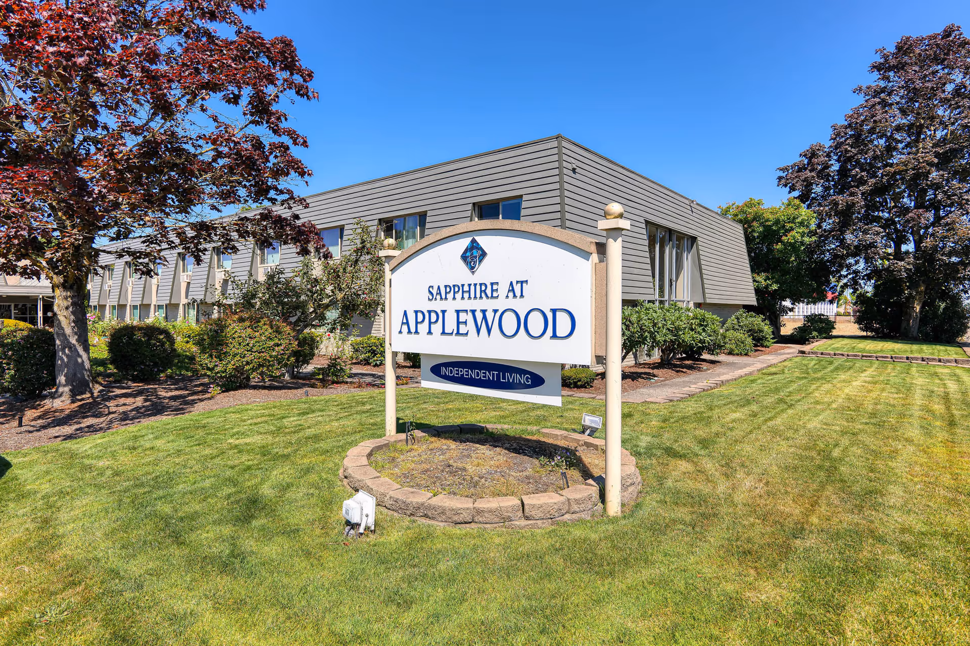 Outdoor view of a senior living facility with a sign that reads 'Sapphire at Applewood Independent Living' in front of a two-story building surrounded by green grass, trees, and shrubs under a clear blue sky.