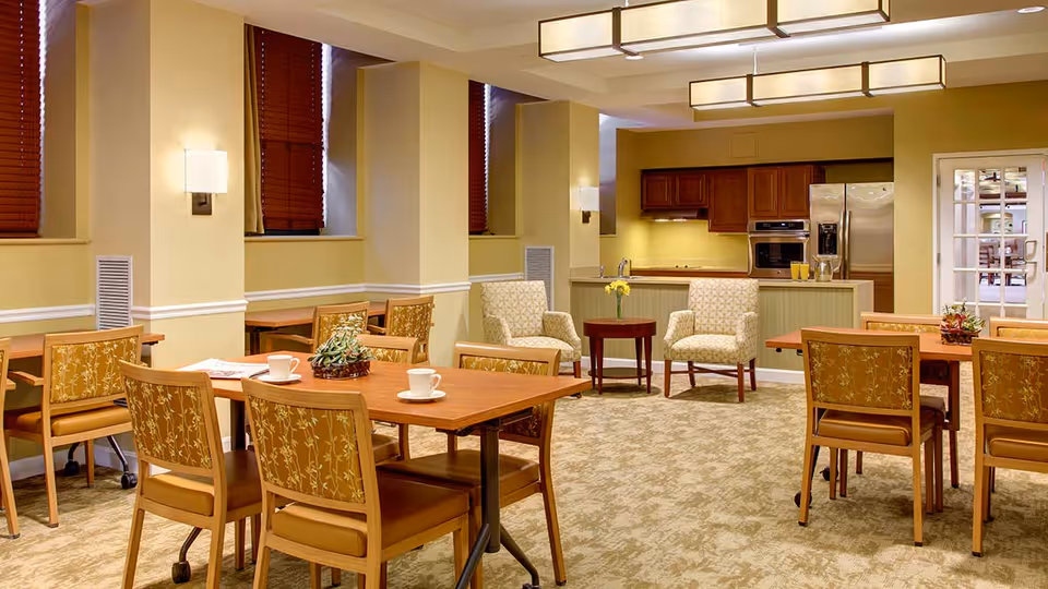 A well-lit dining area with wooden tables and chairs featuring floral upholstery. The room has beige walls with white trim, carpeted floors, and several windows with closed wooden blinds. In the background, there is a kitchen area with wooden cabinets, a stainless steel refrigerator, and a built-in oven. Two upholstered armchairs and a small round table with a vase of flowers are also visible near the kitchen.