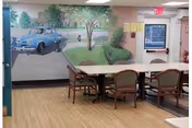A room with a long table surrounded by chairs on a wooden floor. The wall features a mural painting depicting a blue vintage car parked on a driveway with trees and greenery in the background. There is an exit sign above a door and a bulletin board on the wall.