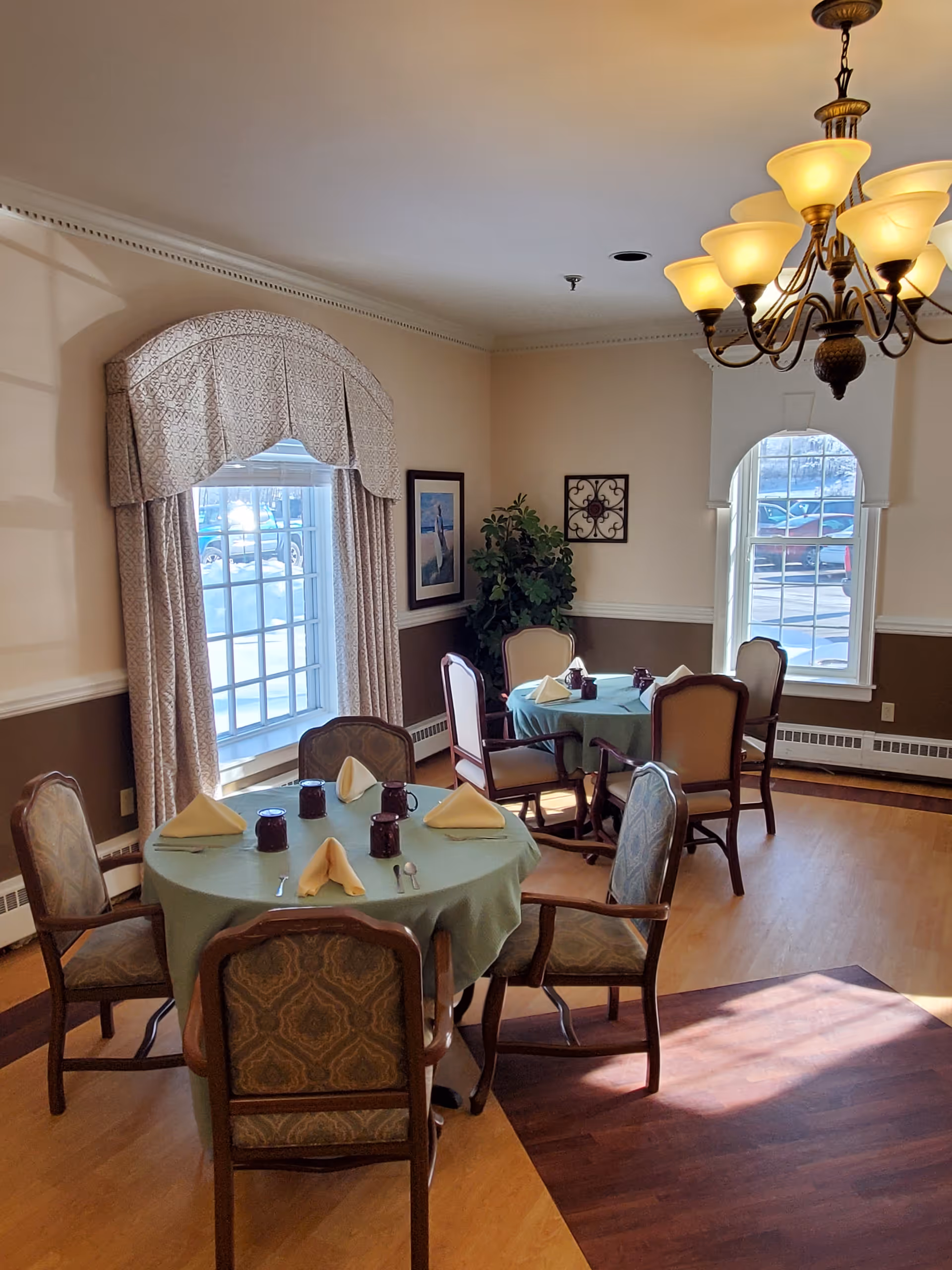 A dining room with two round tables covered with green tablecloths, each set with folded yellow napkins, cups, and silverware. The room has large windows with patterned curtains, a chandelier with multiple lights, framed artwork on the walls, and a potted plant in the corner. The floor is wood, and sunlight streams in through the windows.