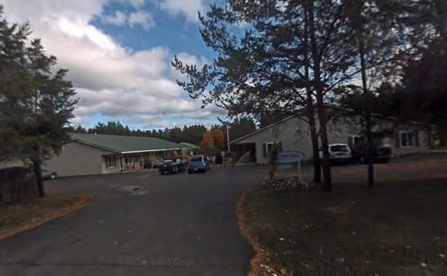 Exterior view of a senior living facility with two single-story buildings surrounded by trees and a paved driveway with parked cars under a partly cloudy sky.