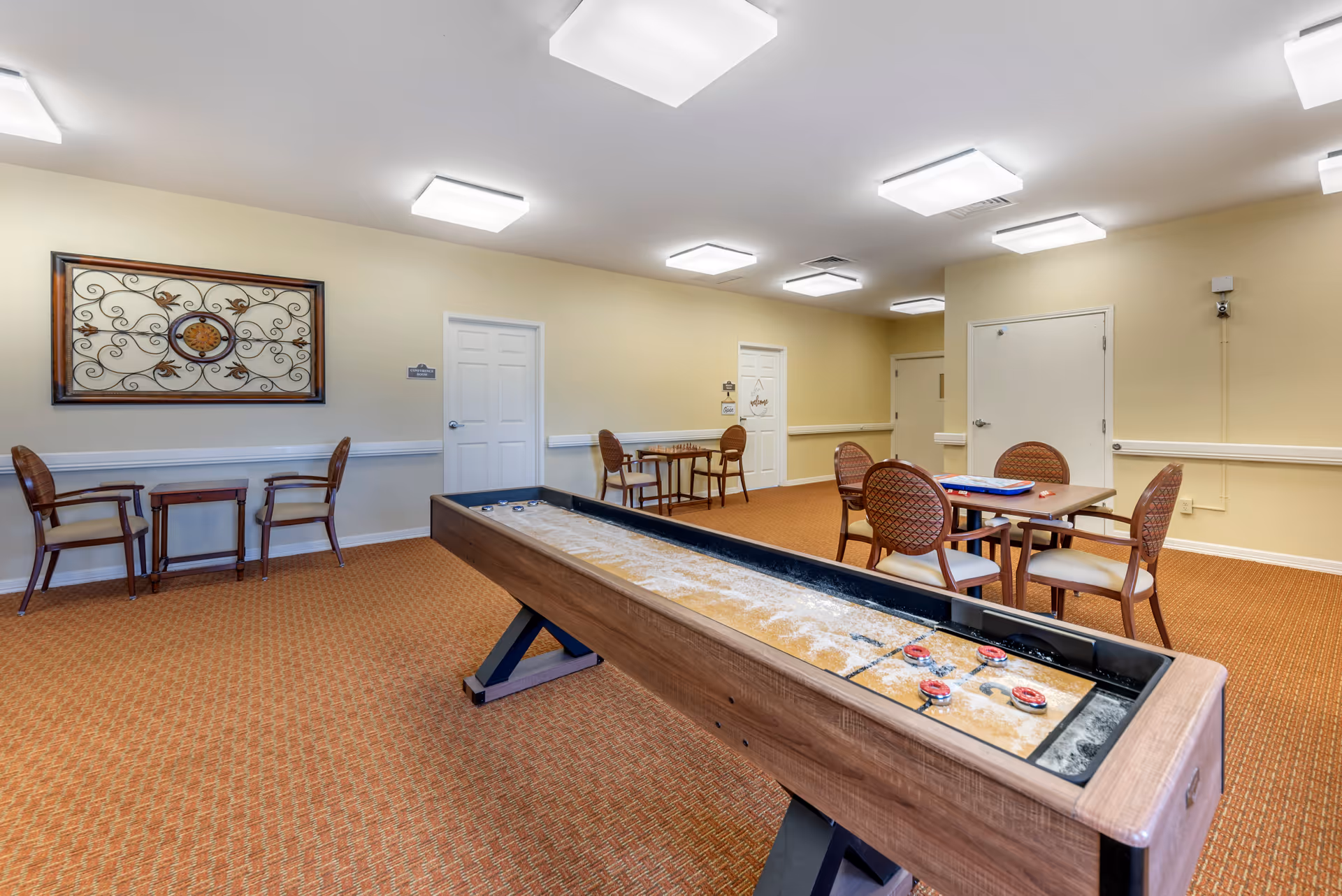 A recreational room with a shuffleboard table in the foreground and several tables with chairs in the background. The room has beige walls, carpeted floor, and ceiling lights. There is decorative wall art on the left wall and multiple closed doors along the back wall.