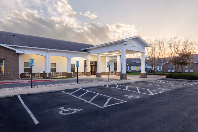 Exterior view of Jackson Creek Memory Care facility at sunset, showing a covered entrance with white columns, brick walls, several windows, and multiple handicap parking spaces in the foreground.
