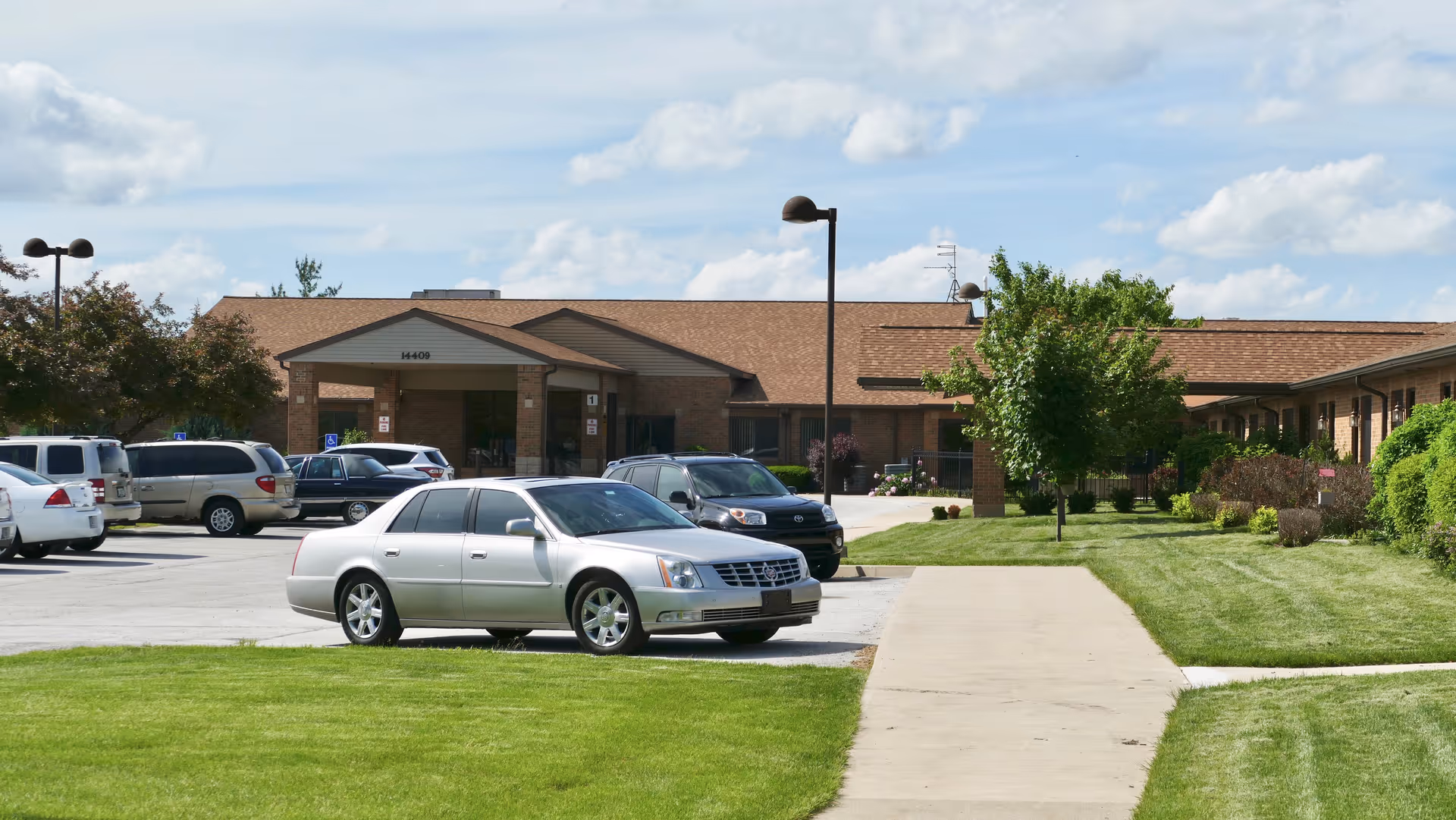 Exterior view of The Cedars senior living facility showing a single-story brick building with a covered entrance, several parked cars in the parking lot, green lawns, trees, and a sidewalk leading to the entrance under a partly cloudy sky.