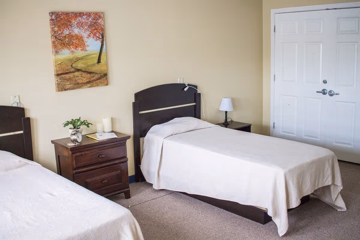 A bedroom with two single beds covered in white bedspreads, a dark wooden nightstand between them holding a small plant, candles, and a book. A painting of a tree with autumn leaves hangs on the beige wall above the nightstand. There is a white double door closet on the right side of the room.