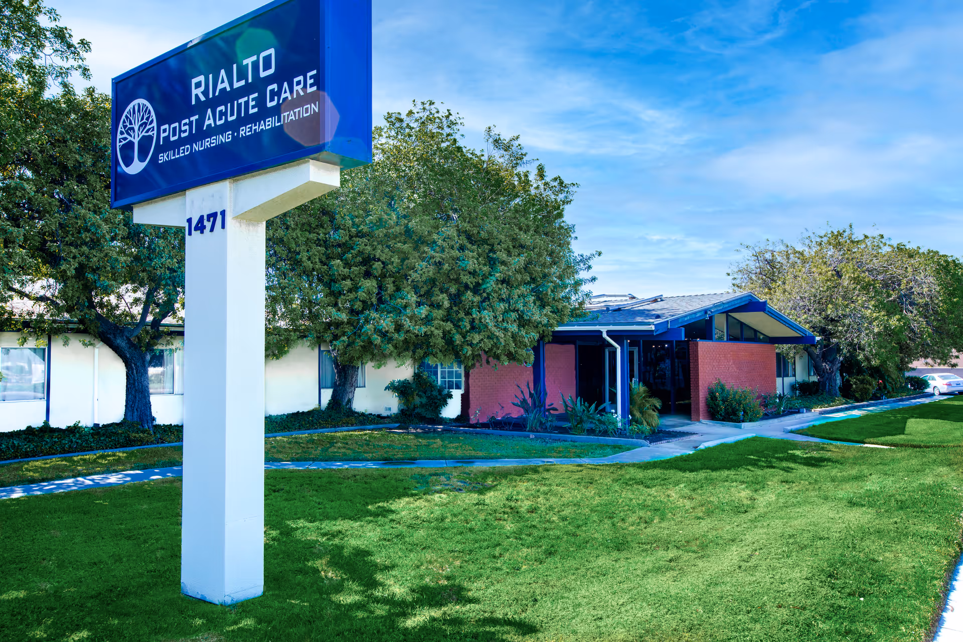 Exterior view of Rialto Post Acute Care facility with a large sign displaying the facility name and services. The building is surrounded by green grass, trees, and a clear blue sky.