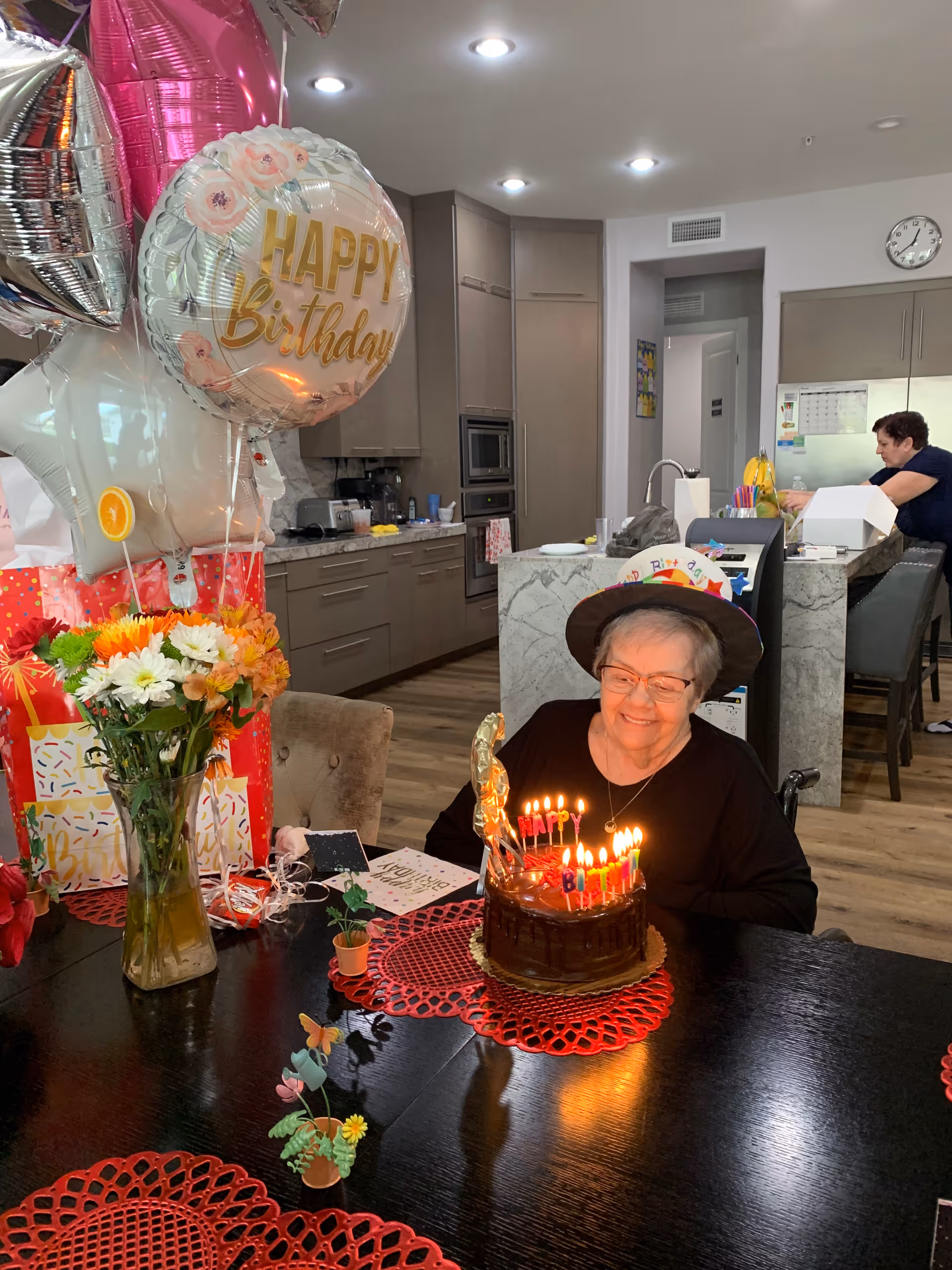 An elderly woman wearing a birthday hat sits at a table with a lit birthday cake in front of her. The table is decorated with red placemats, a vase of colorful flowers, and small decorative plants. Behind her is a modern kitchen with wooden cabinets and a woman working at the counter. There are birthday balloons and wrapped gifts on the left side of the image.