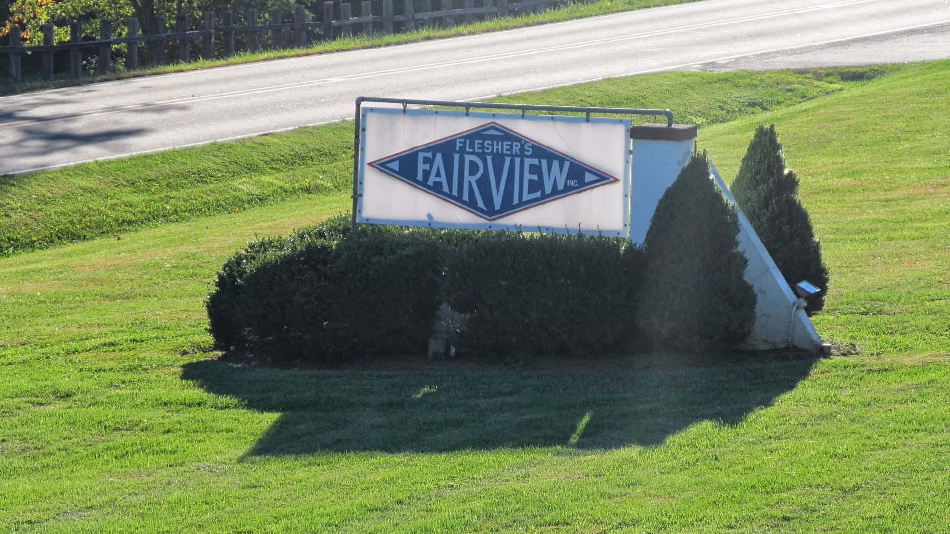A roadside sign for Flesher's Fairview Health Care Center surrounded by green grass and bushes, with a road and guardrail visible in the background.