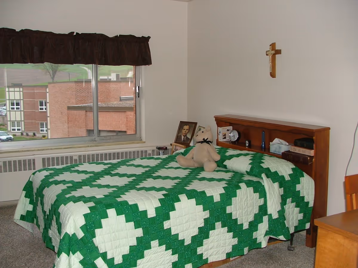A bedroom with a bed covered in a green and white quilt. On the bed is a beige teddy bear with a black ribbon. The headboard has shelves holding books, a clock, a flashlight, and a tissue box. A framed photo is placed on a small table next to the bed. A wooden cross is mounted on the wall above the headboard. A window with a dark valance shows a view of a brick building outside.