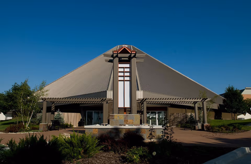 Exterior view of a large pyramid-shaped building with a tall central architectural feature, surrounded by landscaped greenery and benches under a clear blue sky.