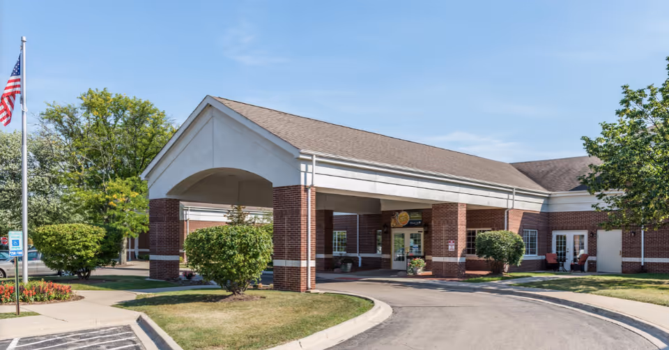 Exterior view of the Citadel of Northbrook facility showing a brick building with a covered entrance, a curved driveway, manicured bushes, an American flag on a flagpole, and a clear blue sky.