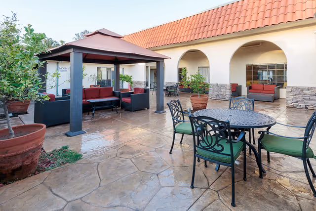 Outdoor patio area with a stone floor, featuring a round metal table with four green cushioned chairs in the foreground. In the background, there is a covered seating area with black wicker furniture and red cushions, large potted plants, and a building with arched doorways and windows.