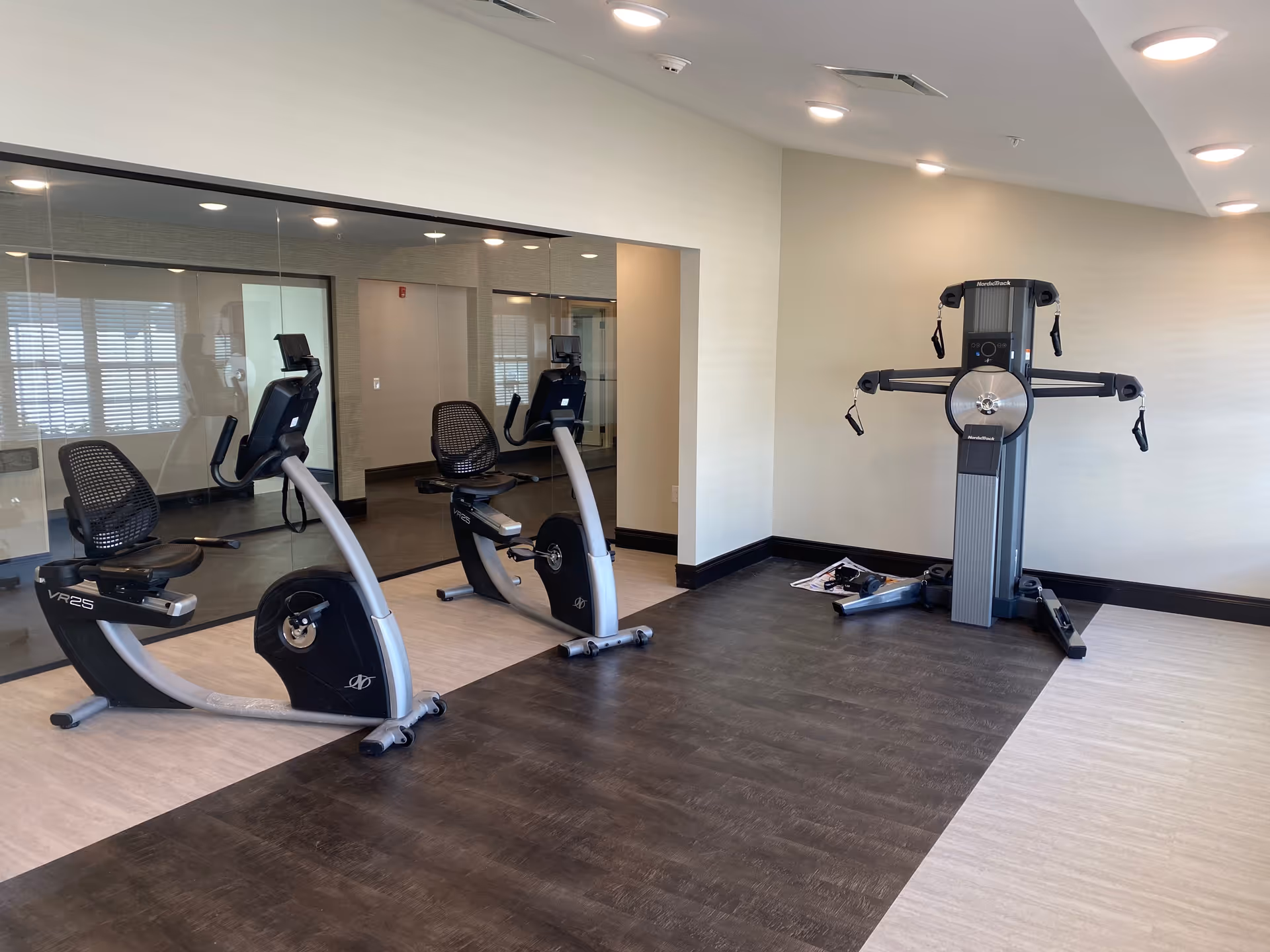 A fitness room with two recumbent exercise bikes positioned side by side on a light-colored floor with a dark wood-like strip in the middle. To the right, there is a multi-functional NordicTrack exercise machine against a beige wall. The room has recessed ceiling lights and a large glass wall reflecting the exercise bikes.
