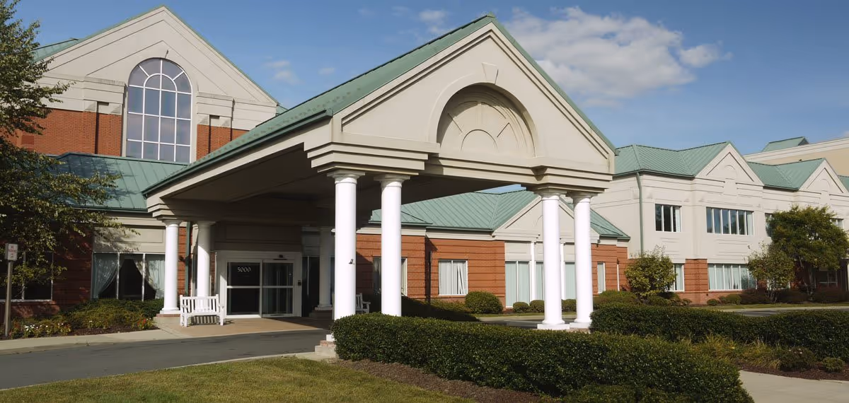 Exterior view of Carnegie Assisted Living At Princeton, showing a large building with a green roof, white columns supporting a covered entrance, and well-maintained landscaping with bushes and trees under a partly cloudy sky.