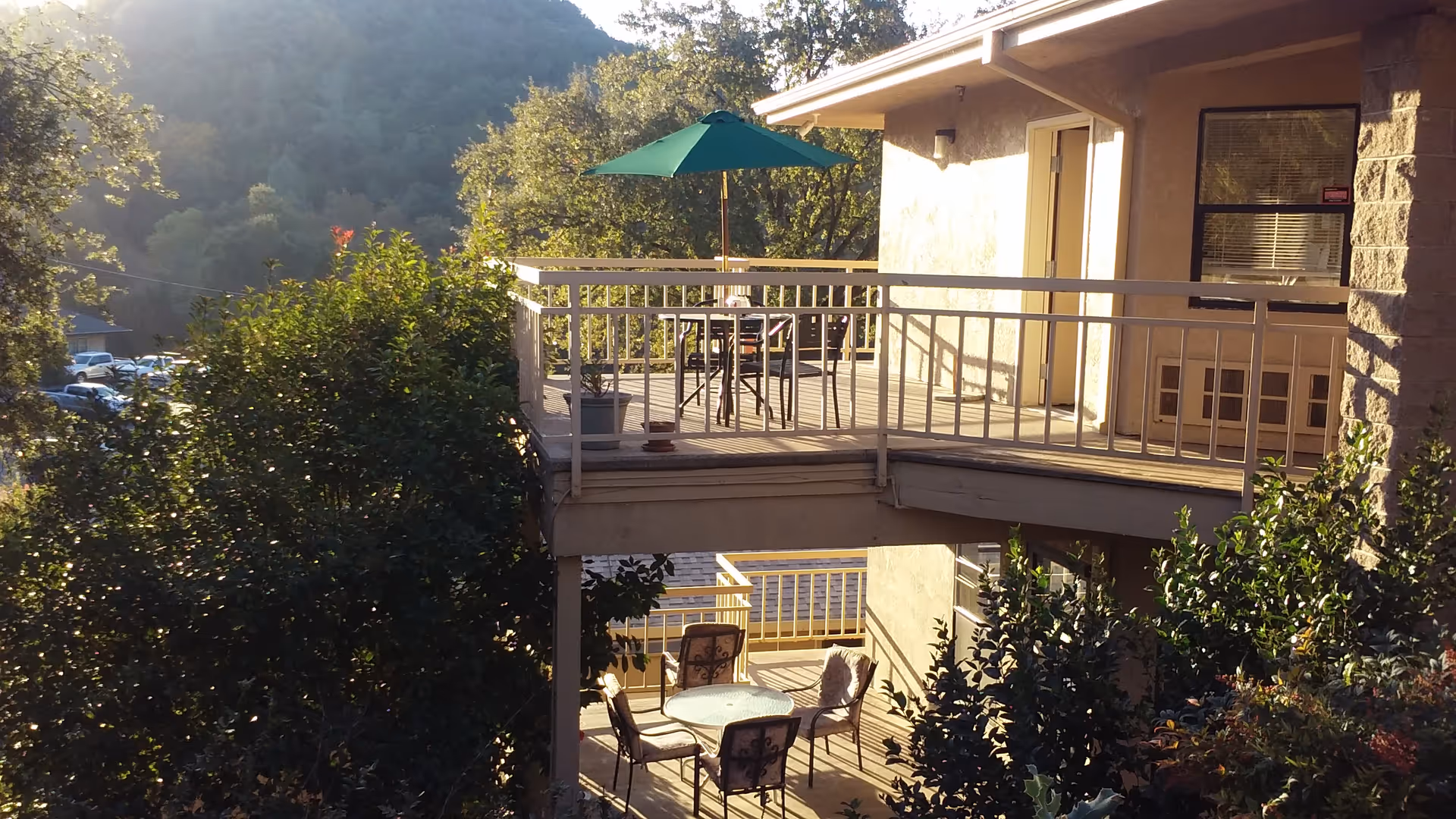 Outdoor view of a senior living facility balcony and patio area with tables, chairs, and a green umbrella surrounded by trees and hills in the background during daylight.
