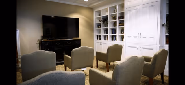 A cozy living room area with five beige upholstered armchairs arranged in a semicircle facing a large flat-screen TV mounted on a dark wooden cabinet. Behind the chairs, there is a white built-in shelving unit with various decorative items and books. The room has soft lighting and a warm, inviting atmosphere.