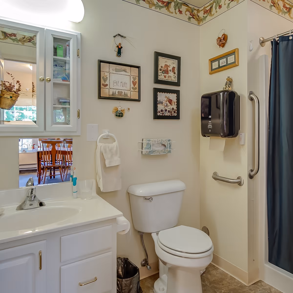 A small bathroom with a white sink and cabinet, a toilet, and a shower with a blue curtain. The walls are decorated with framed pictures and small hanging ornaments. There is a black paper towel dispenser mounted on the wall next to the shower, and a towel ring with a white towel hanging beside the sink. A mirror cabinet is above the sink, reflecting a dining area with wooden chairs and a hanging basket.