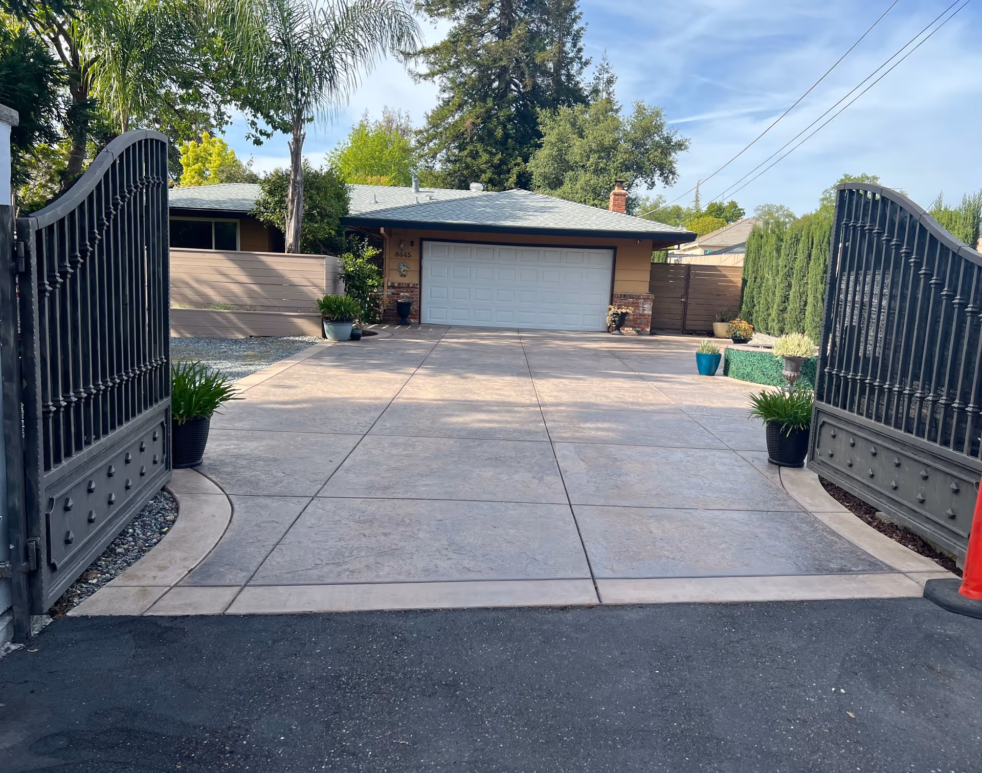 View through open black wrought iron gates onto a wide concrete driveway leading to a single-story house with a two-car garage. The driveway is bordered by potted plants and greenery, with tall trees and shrubs surrounding the property under a partly cloudy sky.
