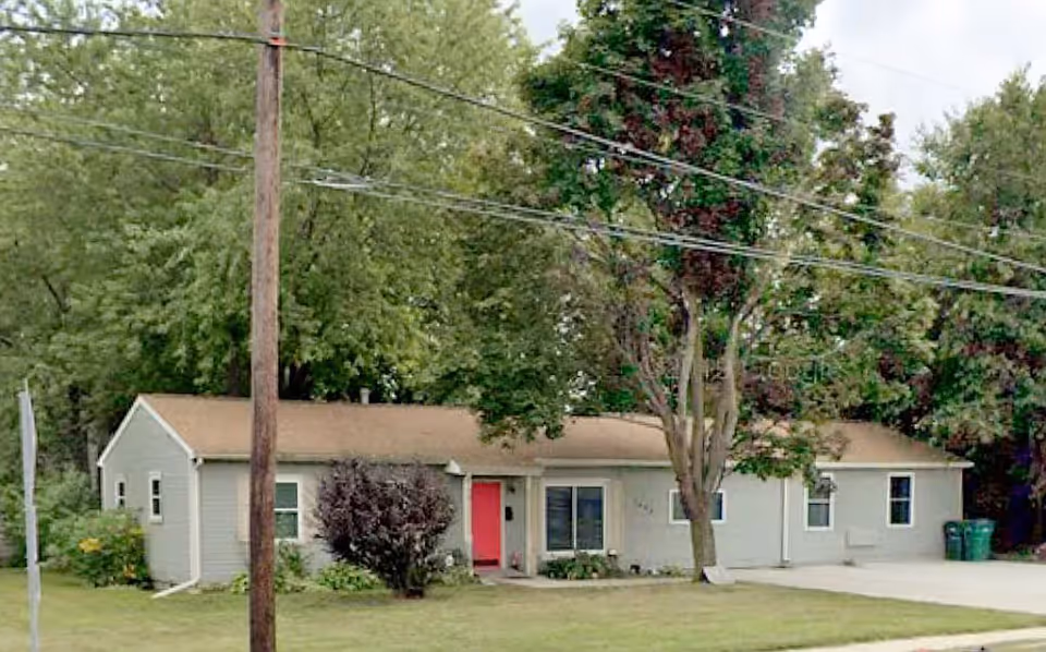 Single-story building with a beige exterior and a brown roof surrounded by trees and greenery. There is a red door at the front entrance and a concrete driveway on the right side. The area is grassy with a few bushes near the building.