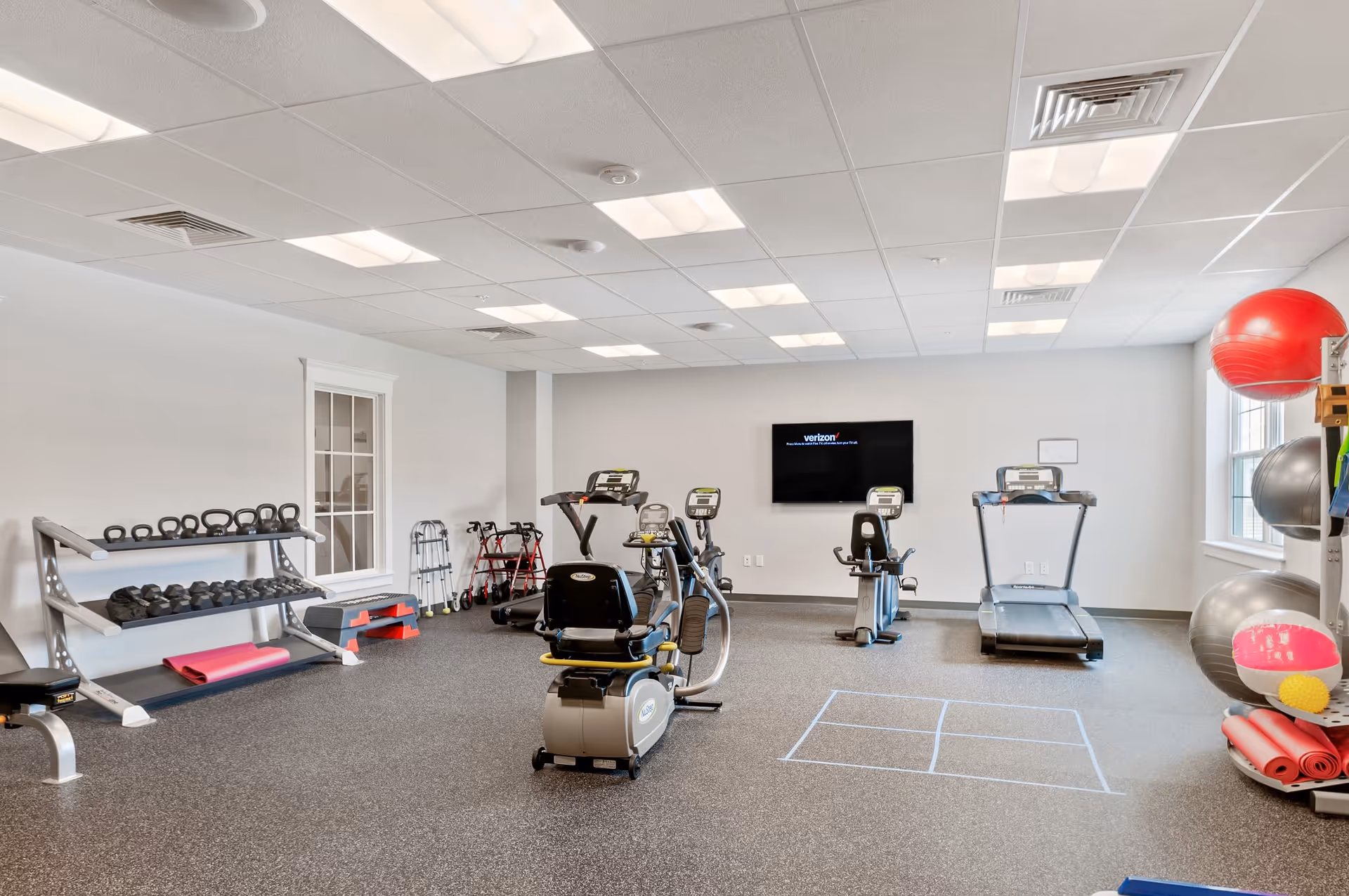 A well-lit fitness room with exercise equipment including treadmills, stationary bikes, kettlebells, dumbbells, exercise balls, and yoga mats. The room has a gray carpeted floor, white walls, and a flat-screen TV mounted on the wall. There are windows letting in natural light and some walkers are visible in the corner.