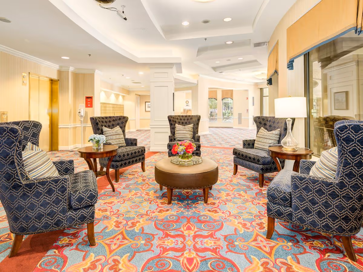 A bright and spacious seating area in a senior living facility with four patterned armchairs arranged around a round ottoman with a flower arrangement on top. There are two small side tables with lamps and flowers. The floor is covered with a colorful patterned carpet, and large windows with yellow valances let in natural light. An elevator and hallway are visible in the background.