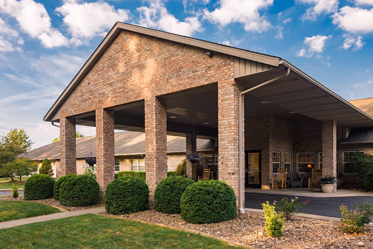 Exterior view of a senior living facility with a large covered entrance supported by brick pillars, surrounded by neatly trimmed bushes and a landscaped garden under a partly cloudy sky.