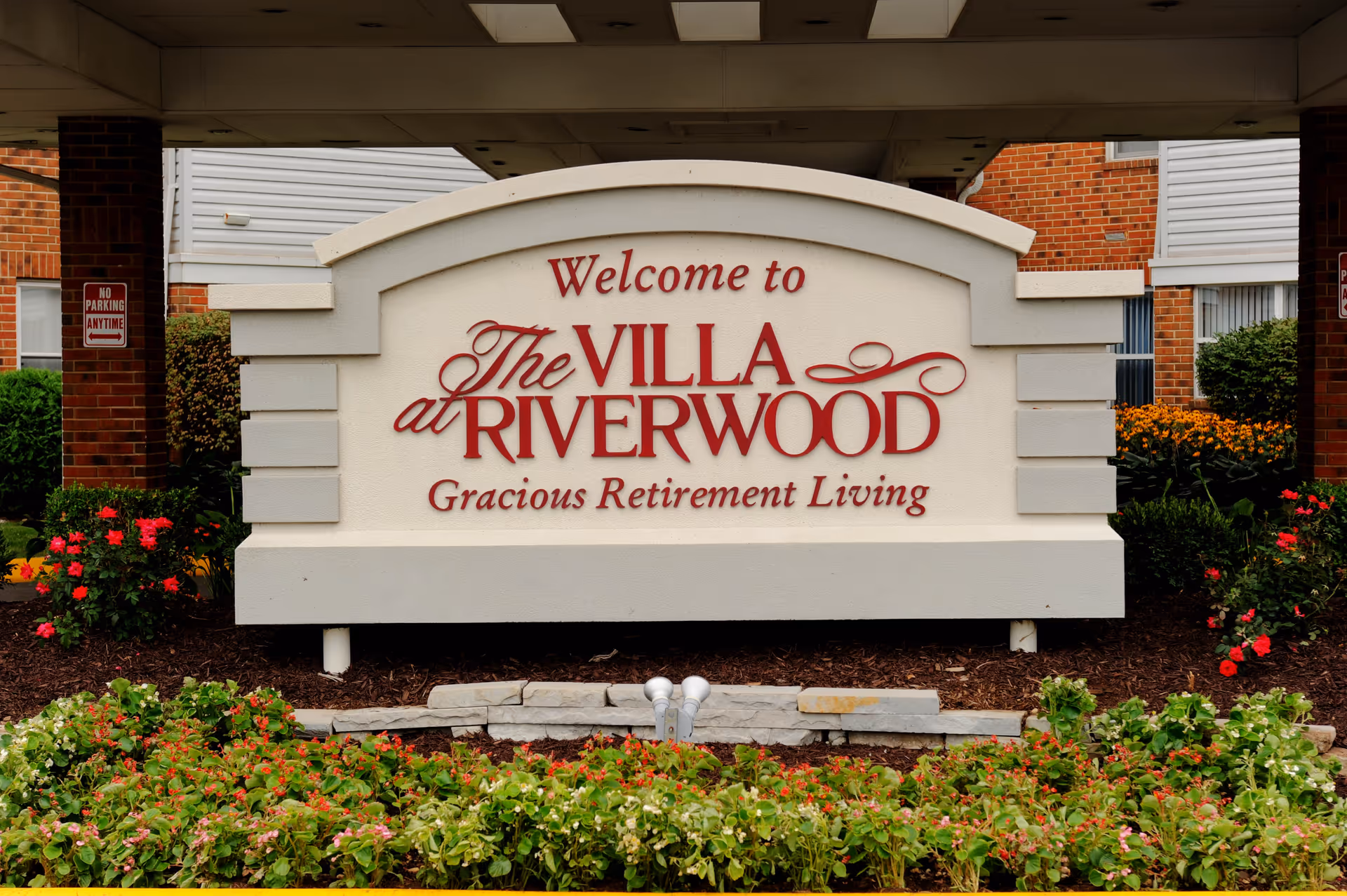 A large welcome sign for The Villa at Riverwood, a gracious retirement living facility, surrounded by flowers and greenery with parts of the building visible in the background.