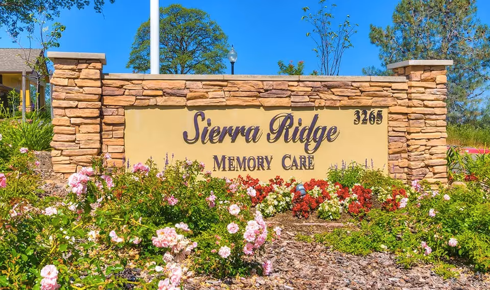 Stone and stucco sign for Sierra Ridge Memory Care surrounded by colorful flowers and greenery under a clear blue sky.