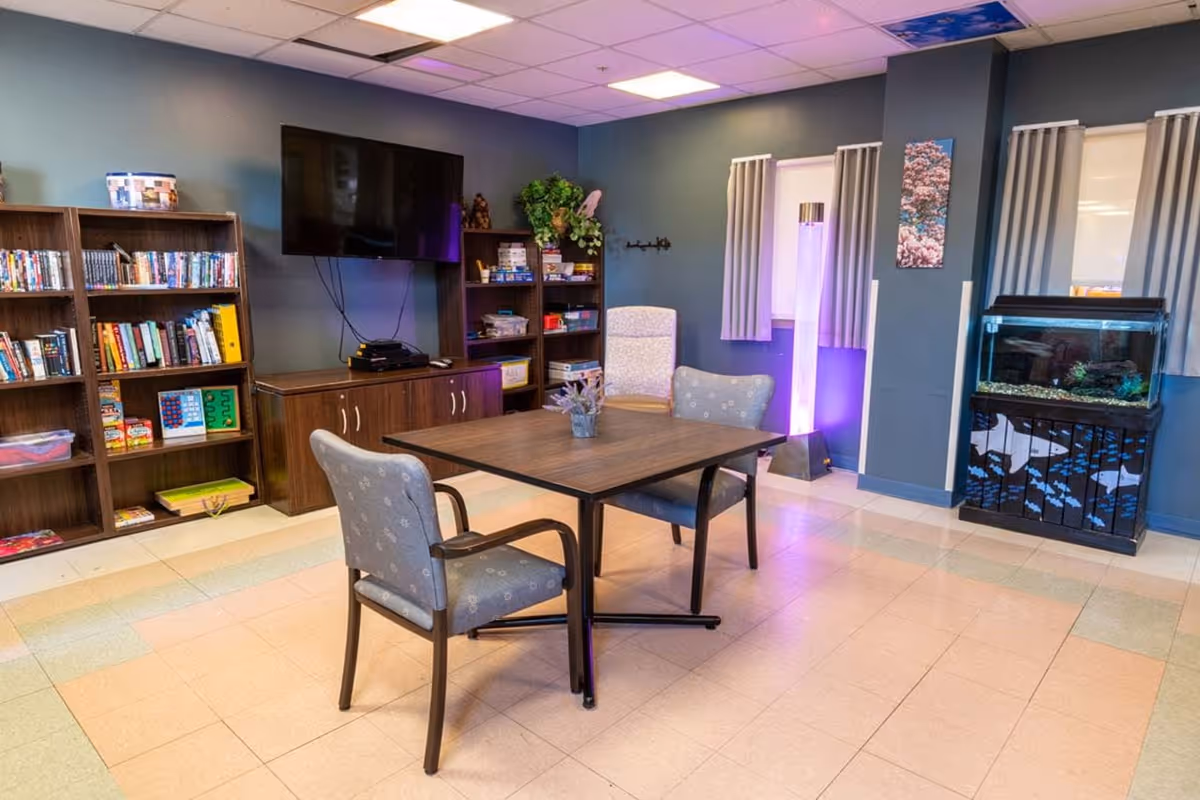 A cozy common room with a square wooden table and two cushioned chairs in the center. The room features bookshelves filled with books and games, a wall-mounted flat-screen TV, a tall illuminated lamp with purple light, and a fish tank on a decorated stand. The walls are painted blue with two small windows covered by curtains.
