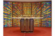 Interior view of a chapel or meditation room featuring a large, colorful stained glass window with geometric patterns in red, blue, yellow, and green hues. In front of the window is a wooden altar or podium on a red carpeted floor.