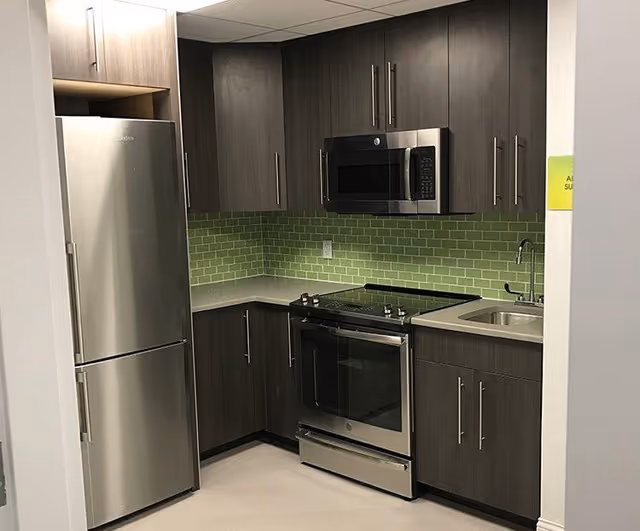 Modern kitchen area with stainless steel refrigerator, oven, and microwave. The kitchen features dark wood cabinets, a green tiled backsplash, a countertop with a sink, and a light-colored floor.