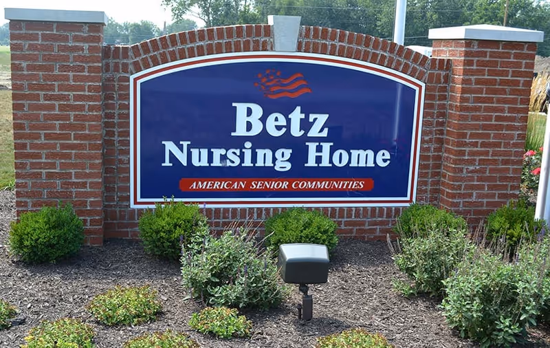 A large brick sign for Betz Nursing Home with a blue background and white text, featuring a small American flag graphic and the phrase 'American Senior Communities' below the name. The sign is surrounded by landscaping with green bushes and mulch.