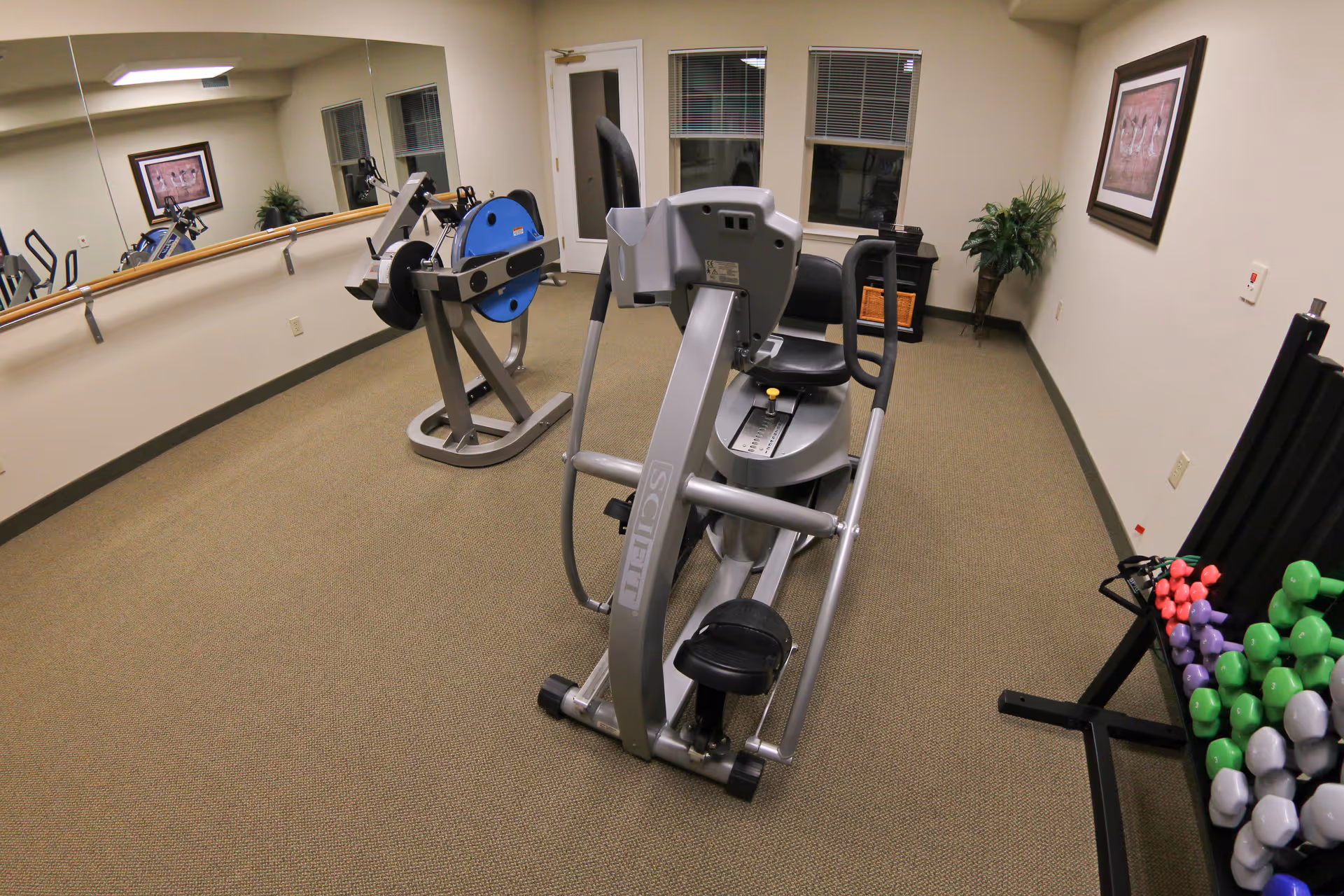 A small fitness room with exercise equipment including a recumbent elliptical machine, a hand cycle machine, and a rack of colorful dumbbells. The room has beige walls, a large mirror on one side, a framed picture on the wall, and a potted plant in the corner.