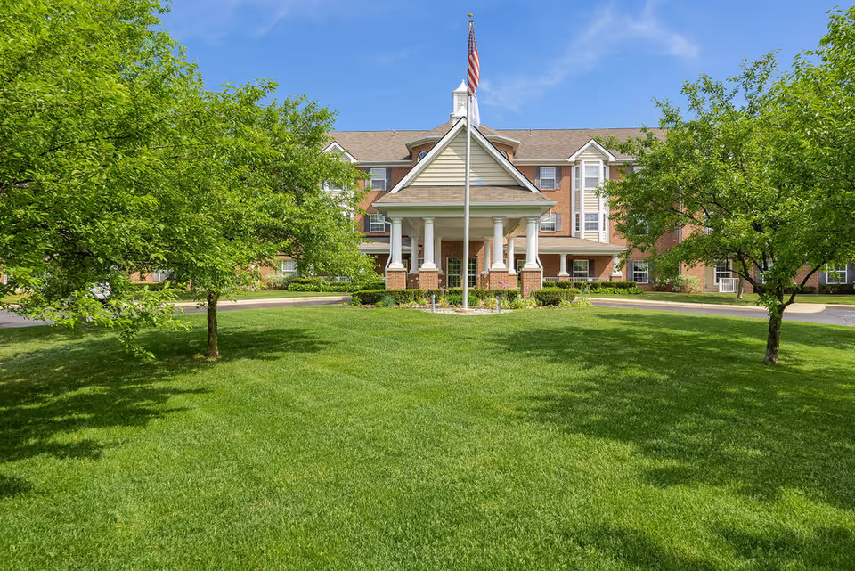 Front exterior view of Elmhaven Manor, a multi-story brick building with a covered entrance supported by white columns, an American flag on a flagpole in front, surrounded by green grass and trees under a clear blue sky.