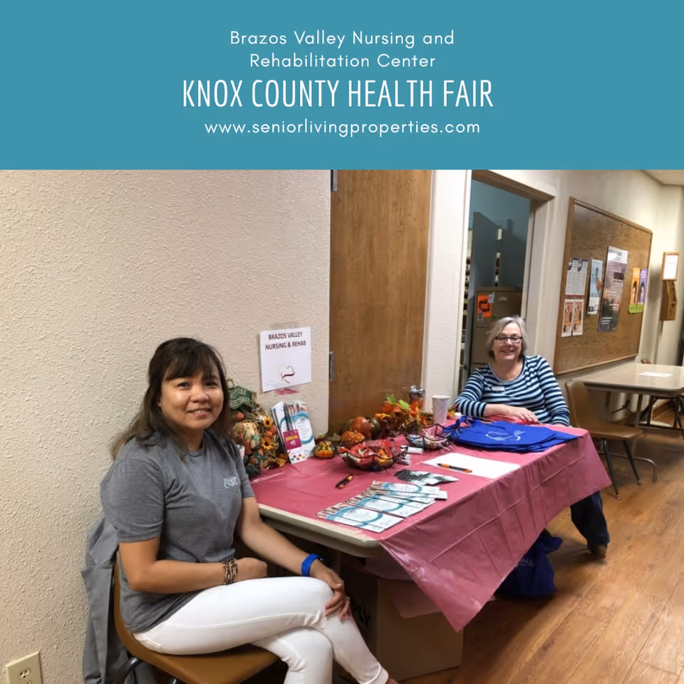 Two women sitting at a table covered with a pink tablecloth inside a hallway. The table has brochures, pens, and small decorative pumpkins. A sign on the wall behind them reads 'Brazos Valley Nursing & Rehab'. The hallway has wooden floors and bulletin boards on the walls.