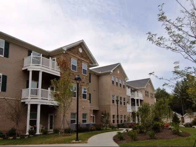 Exterior view of a multi-story senior living facility building with balconies, windows, and a landscaped walkway surrounded by trees and shrubs under a partly cloudy sky.