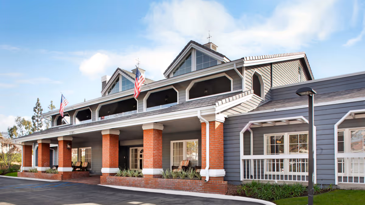 Exterior view of a senior living facility building with gray siding, red brick columns, and white trim. The building features a covered porch with seating and American flags displayed on the roof. The sky is partly cloudy.