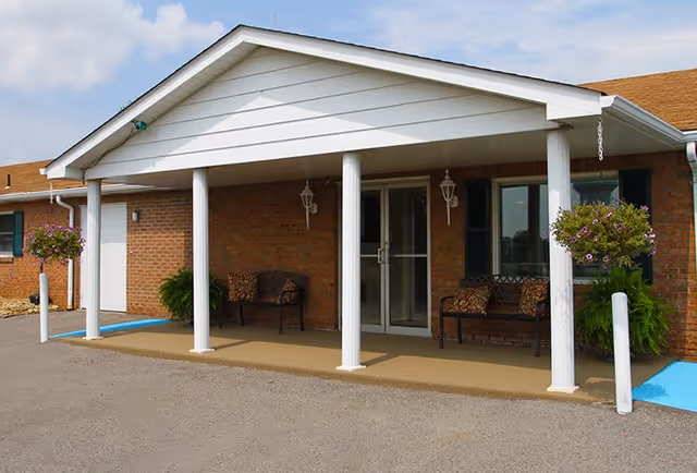 Entrance of a brick building with a white covered porch supported by four white columns. There are two benches with patterned cushions under the porch, two hanging flower baskets on either side, and double glass doors in the center. The ground is paved with parking spaces marked in blue nearby.