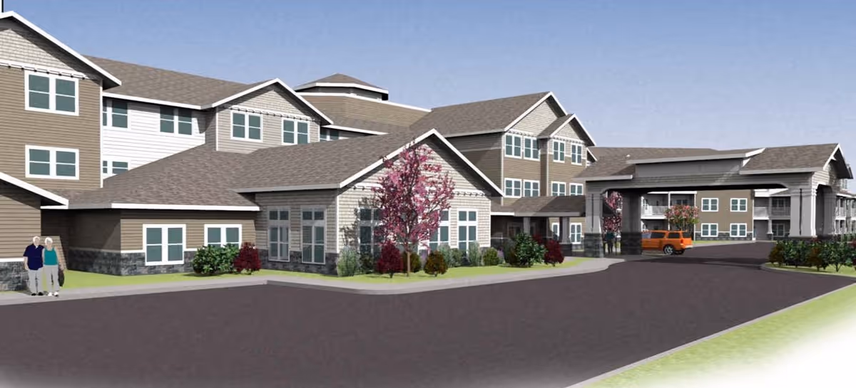 Exterior view of a multi-story senior living facility with beige and white siding, multiple windows, and a covered entrance. There are landscaped bushes and trees with pink blossoms near the building. A couple of elderly people are walking on the sidewalk, and an orange vehicle is parked near the entrance under the covered area. The sky is clear and blue.