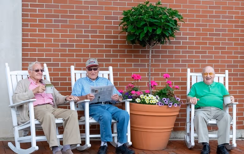Three elderly men sitting on white rocking chairs outside against a brick wall. One man is holding a newspaper, and the other two are holding mugs. There is a large flower pot with colorful flowers and a small tree between them.