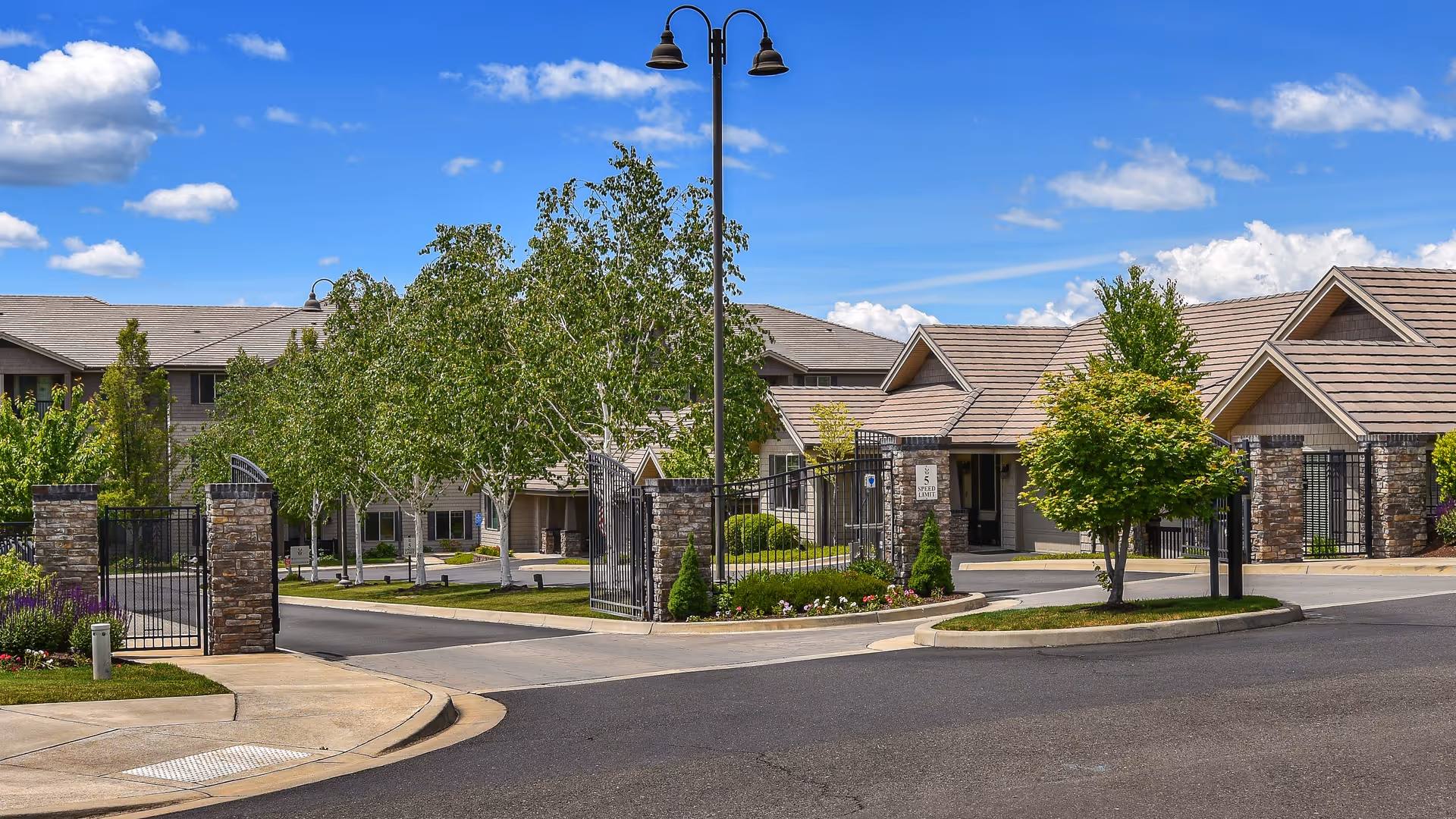 Gated entrance to a senior living facility with stone pillars, black metal gates, landscaped greenery, and a clear blue sky with some clouds.
