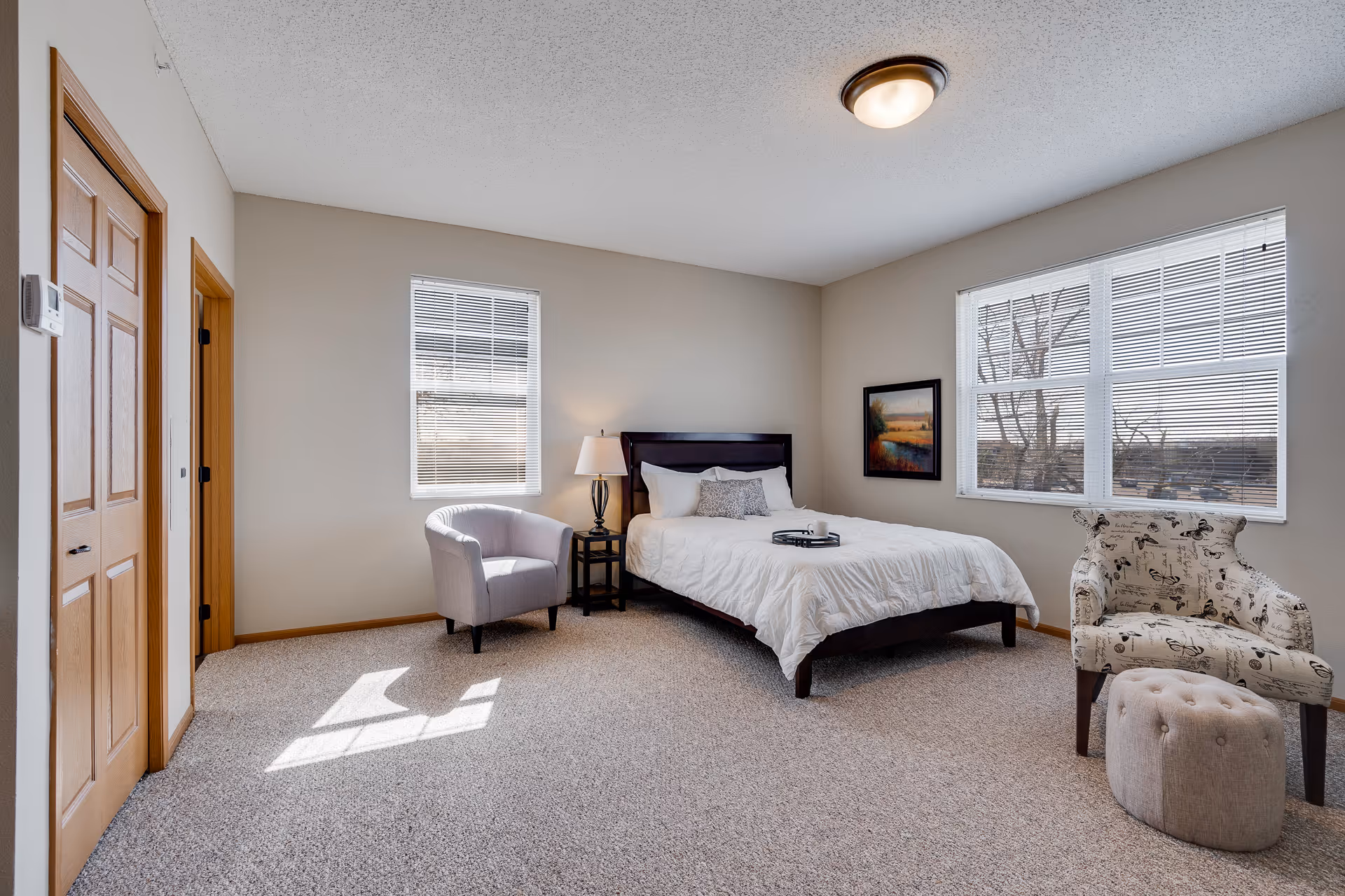 A bright bedroom with a large window, a bed with white bedding and pillows, a nightstand with a lamp, a gray armchair, and a patterned armchair with a matching ottoman on a carpeted floor.