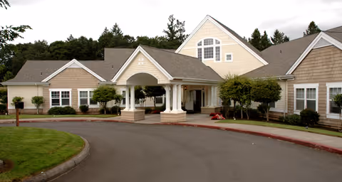 Exterior view of a single-story senior living facility building with beige siding, white trim, and a covered entrance. The driveway curves in front of the entrance, with green lawns and trees surrounding the building under a cloudy sky.