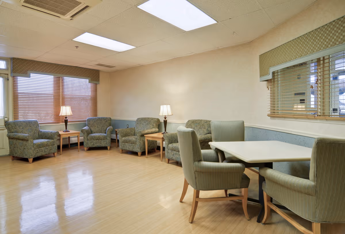 A well-lit common room with four upholstered armchairs arranged around two wooden side tables with lamps, and a square table with four cushioned chairs. The room has large windows with wooden blinds and light-colored walls with a blue wainscoting.