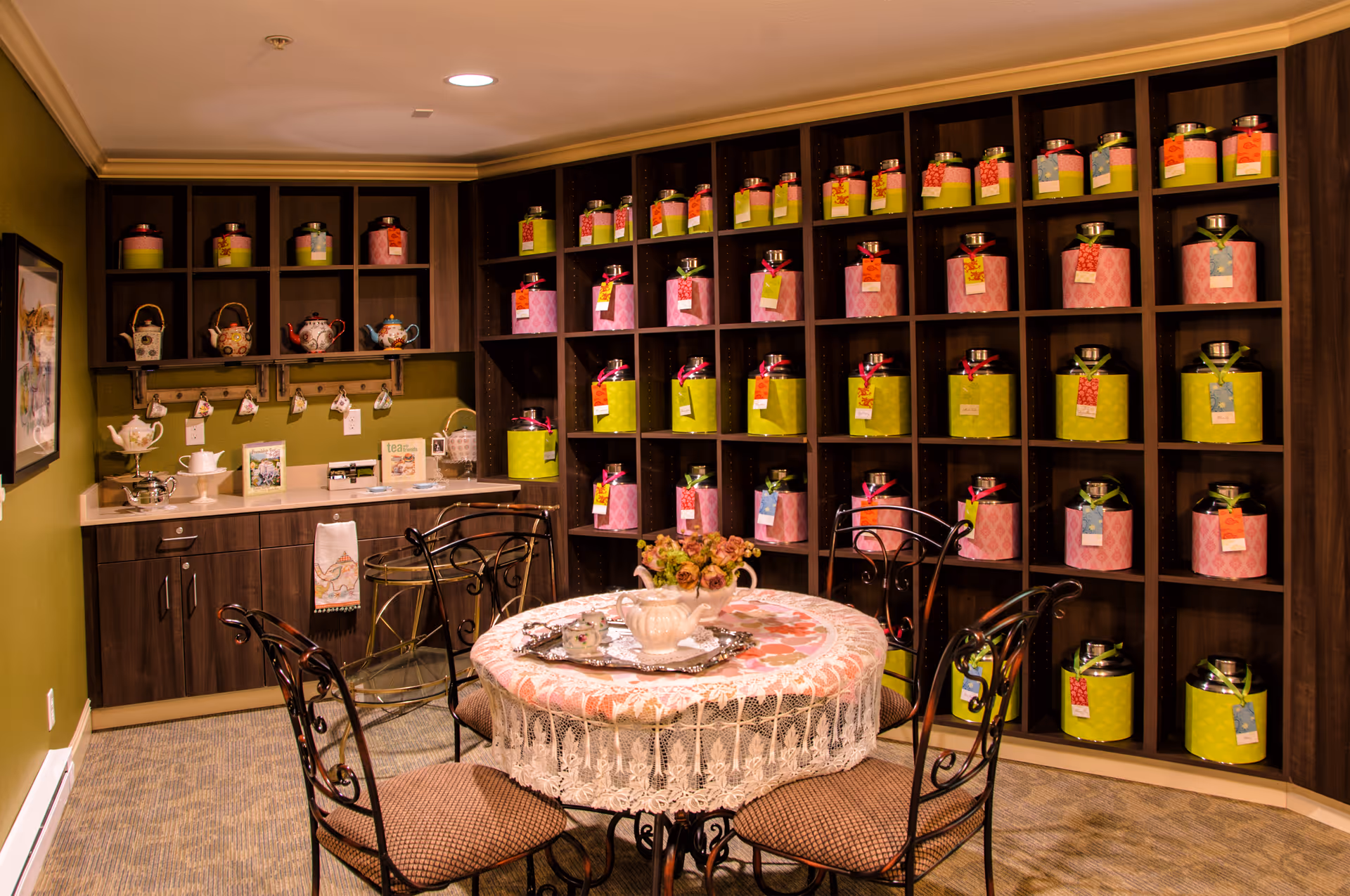 A cozy tea room with a round table covered by a lace tablecloth and a floral centerpiece. Surrounding the table are four wrought iron chairs with cushioned seats. The walls are lined with dark wooden shelves filled with colorful tea canisters and teapots. A small counter with cabinets and hanging teacups is visible on the left side of the room.